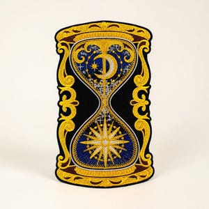 May include: Embroidered patch with an hourglass design and celestial details. The patch has a black background with gold and blue accents. The top half features a crescent moon and star, while the bottom half has a starburst.