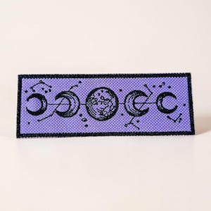 May include: A rectangular patch with a celestial design. It has a lavender background and a black border. The design features phases of the moon and constellations in black. The patch is approximately 10 cm long and 4 cm wide.