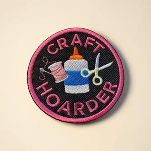 Pode incluir: Patch bordado circular com as palavras "CRAFT HOARDER" em rosa. O design inclui um frasco de cola, tesouras e uma bobina de linha. O patch tem um fundo preto com uma borda rosa, ideal para artesãos.