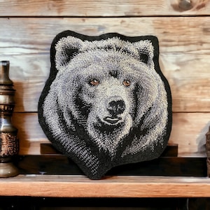 Op de afbeelding: Gestikt zwart kussen met een gedetailleerd beeld van de kop van een bruine beer.