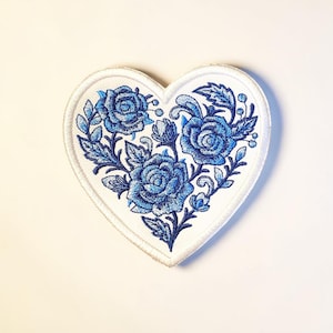 May include: A heart-shaped embroidered patch featuring blue roses and foliage on a white background. The patch has a white border and is likely designed to be sewn or ironed onto fabric. The floral design is detailed and intricate.