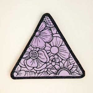 May include: A triangular patch featuring a floral design. The patch has a light purple background with black outlines of various flowers. The patch is bordered with a black, glittery trim.