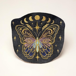 Puede incluir: Parche bordado con un diseño de mariposa y elementos celestiales. La mariposa tiene detalles morados, azules y dorados sobre un fondo negro. El diseño incluye fases lunares, estrellas y detalles florales.