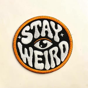 May include: A circular embroidered patch with a black background and an orange border. The text STAY WEIRD is stitched in white with a stylized eye icon positioned between the two words. Two small white four-pointed stars flank the eye symbol.