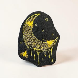 May include: A black felt patch featuring a crescent moon design with yellow embroidery. The moon has a honeycomb pattern and dripping details. Small stars and two moth-like figures are also embroidered on the patch.