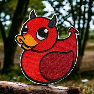 May include: Red embroidered patch of a cartoon devil duck with large eyes and horns. The duck is sitting on a brown log.