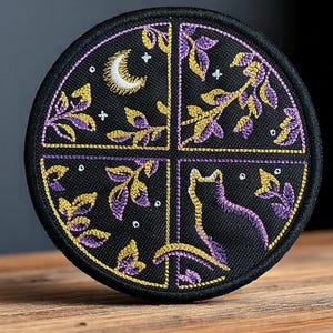May include: Round black embroidered patch featuring a cat silhouette, crescent moon, and stylized leaves. The design incorporates gold and purple thread. The patch is divided into four sections with a gold border and white accent dots.
