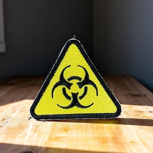 May include: A yellow triangular patch featuring a black biohazard symbol, with a black border. The patch is placed on a wooden surface. The biohazard symbol is a warning sign for biological hazards.