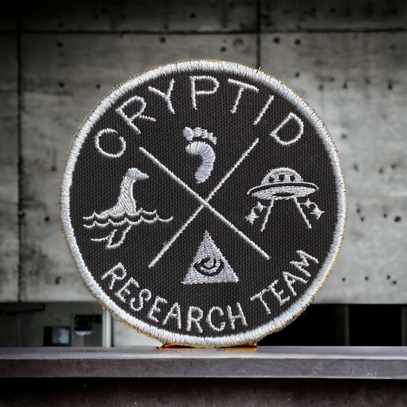 May include: A black and white embroidered patch with the text "Cryptid Research Team" in a circle. The patch features a footprint, a UFO, a sea monster, and an all-seeing eye.