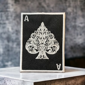 Könnte beinhalten: Schwarzer Pik-Ass-Aufnäher mit detailliertem weißem Pik-Design und dem Buchstaben "A" in den Ecken. Der Aufnäher hat einen weißen Rand und wird auf einer weißen Oberfläche präsentiert.