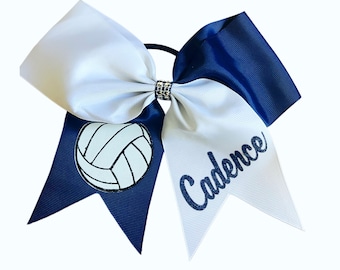 May include: A white and navy blue cheer bow with a white volleyball graphic and the name "Cadence" in glitter lettering.