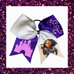 May include: Purple and white cheer bow with a pink glitter castle and a character from the movie "Encanto" holding a cupcake. The bow has a purple ribbon with a pink glitter swirl design.