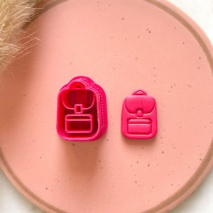 Może przedstawiać: Dwa jasnoróżowe wykrawacze do ciastek w kształcie plecaka. Jeden jest większy od drugiego. Wykrawacze znajdują się na różowym talerzu z brązową obwódką. W tle suszona trawa ozdobna.