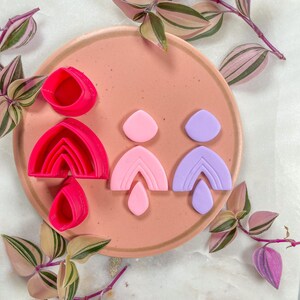 Może przedstawiać: Zestaw elementów i wykrawaczy do kolczyków z gliny polimerowej w odcieniach różu i fioletu, ułożonych na talerzu w kolorze brzoskwiniowym. Elementy obejmują kształty łuków i kropli, gotowe do tworzenia. Zielone i różowe gałązki roślin otaczają talerz.