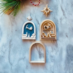 May include: Three cookie cutters in the shape of a Christmas village scene. The cutters are made of beige plastic and feature a star, a house with a Christmas tree, and a house with Santa Claus flying overhead.