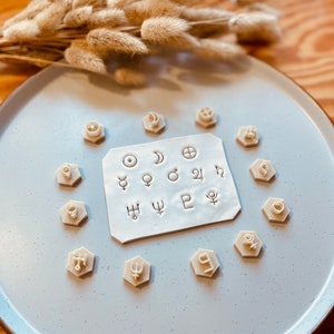 Peut inclure: Un ensemble de 12 tampons astrologiques en forme hexagonale de couleur crème. Les tampons sont disposés autour d'une carte blanche sur laquelle sont imprimés les mêmes symboles. La carte est sur une assiette bleu clair.