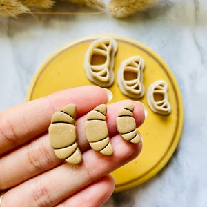 May include: Three different sized croissant shaped clay earrings in a light brown color. The earrings are displayed on a yellow plate with other croissant shaped earrings.