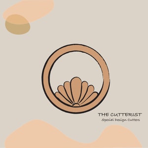 May include: A tan-coloured cookie cutter featuring a circular frame and a stylised seashell design at the bottom. The text "THE CUTTERIST Special Design Cutters" is printed in black below the cutter.