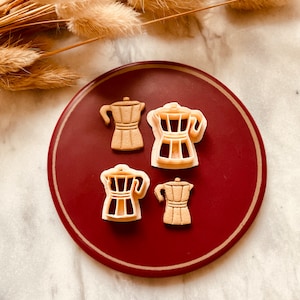Könnte beinhalten: Vier braune Plastik-Ausstecher in Form von Kaffeekannen auf einem roten Teller. Der Teller liegt auf einer weißen Marmorfläche.