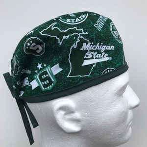 May include: Green surgical scrub hat with a white Michigan State logo and various other white logos and designs. The hat has two green ties.