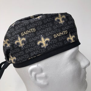 May include: Black and gold surgical scrub hat with a repeating pattern of the New Orleans Saints logo and the word "Saints".