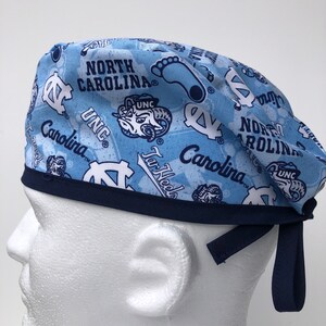 Puede incluir: Un gorro de quir&oacute;fano azul y azul marino con un patr&oacute;n repetido del logotipo de los Tar Heels de la Universidad de Carolina del Norte, las palabras "North Carolina", "UNC" y "Tar Heels", y una silueta de una huella. El gorro tiene una banda el&aacute;stica azul marino y lazos.