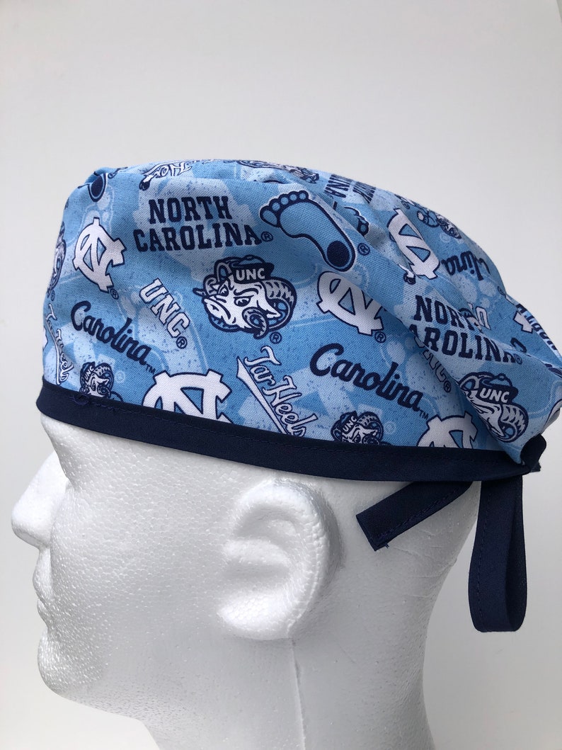 Puede incluir: Un gorro de quir&oacute;fano azul y azul marino con un patr&oacute;n repetido del logotipo de los Tar Heels de la Universidad de Carolina del Norte, las palabras "North Carolina", "UNC" y "Tar Heels", y una silueta de una huella. El gorro tiene una banda el&aacute;stica azul marino y lazos.