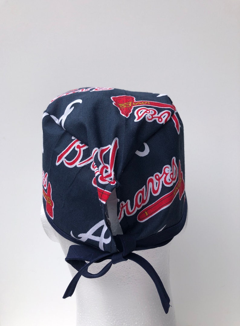 Peut inclure: Un bonnet de chirurgien bleu et rouge avec un motif r&eacute;p&eacute;t&eacute; du logo des Braves d'Atlanta. Le bonnet a une cravate &agrave; l'arri&egrave;re.