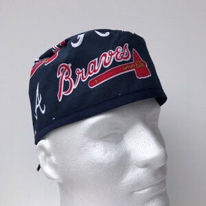 Peut inclure: Un bonnet de chirurgien bleu et rouge avec le logo des Braves d'Atlanta et le mot "Braves" en lettres blanches.