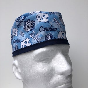 Puede incluir: Un gorro de quir&oacute;fano azul claro con ribete azul marino. El gorro est&aacute; cubierto con un patr&oacute;n del logotipo de los Tar Heels de la Universidad de Carolina del Norte, las palabras "UNC", "Carolina" y "Tar Heels", y la mascota de la escuela, el carnero.