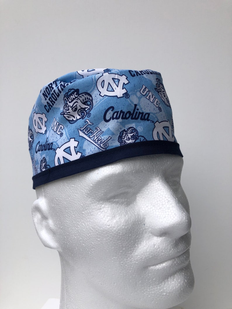 Puede incluir: Un gorro de quir&oacute;fano azul claro con ribete azul marino. El gorro est&aacute; cubierto con un patr&oacute;n del logotipo de los Tar Heels de la Universidad de Carolina del Norte, las palabras "UNC", "Carolina" y "Tar Heels", y la mascota de la escuela, el carnero.