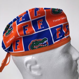 May include: A blue, orange, and white patterned scrub cap featuring the Florida Gators logo and text. The cap has a repeating pattern of the letter "F" and the team's mascot, an alligator. The cap is tied at the back with orange ties.