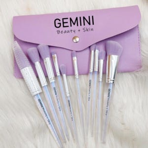 Könnte beinhalten: Ein Set mit 10 Make-up-Pinseln mit durchsichtigen Griffen und violetten Borsten. Die Pinsel sind in einer Reihe angeordnet und mit "GEMINI Beauty + Skin" beschriftet. Die Pinsel befinden sich in einer violetten Tasche.