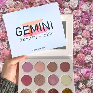 Może przedstawiać: Biała paleta cieni do powiek z 12 odcieniami. Paleta ma białe tło z czarną etykietą, na której widnieje napis "GEMINI Beauty + Skin 12 SHADE PALLETE".