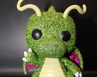 May include: Green glitter dragon figurine with pink wings and yellow horns. The figurine has black eyes and a yellow belly.