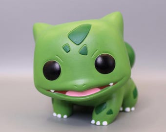 May include: A green Bulbasaur figure with large black eyes, a smiling mouth, and white toes. The figure has a bulb on its back and a leaf-shaped marking on its head. The figure is a collectible item.