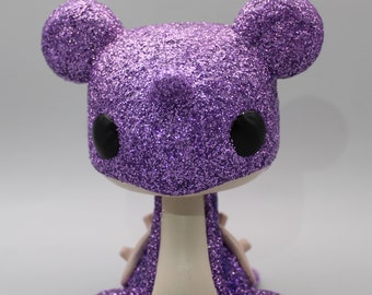 May include: Purple glitter dragon figurine with black eyes and a white belly. The dragon is sitting on its hind legs with its front legs tucked in.