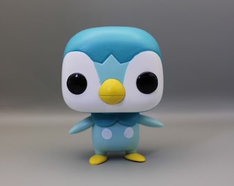 May include: A blue and white penguin figurine with a yellow beak and black eyes. The figurine is standing on a gray surface.