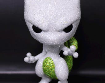May include: A white and green glitter figurine of a Pokemon character, Mewtwo. The figurine is standing on a clear plastic base.