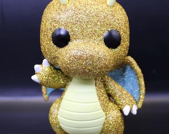 May include: A gold glitter Dragonite Funko Pop figure with a light green body and blue glitter wings. The figure has black eyes and yellow antennas.