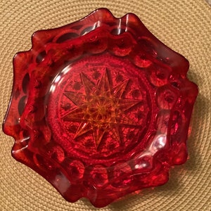 May include: A red glass ashtray with a starburst design. The ashtray has a scalloped edge and is made of clear glass with a red tint.