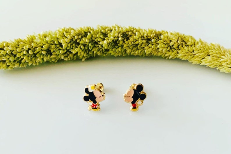 Puede incluir: Un par de pendientes de tono dorado con una imagen de dibujos animados de Mickey Mouse y Minnie Mouse bes&aacute;ndose. Los pendientes son peque&ntilde;os y delicados.
