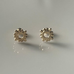 May include: A pair of gold-toned earrings with a sun-like design. Each earring features a circular center with a textured, sunburst-like pattern.