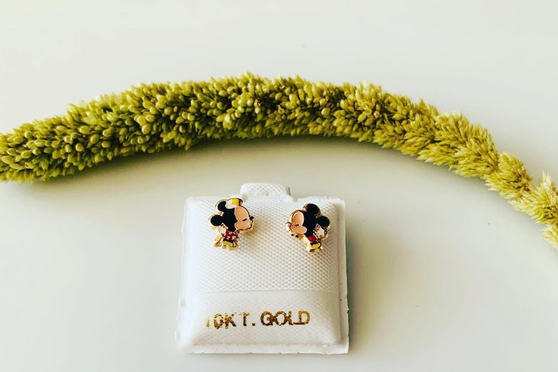 Puede incluir: Un par de pendientes de oro con Mickey Mouse y Minnie Mouse en una tarjeta de presentaci&oacute;n blanca con el texto "10KT GOLD".