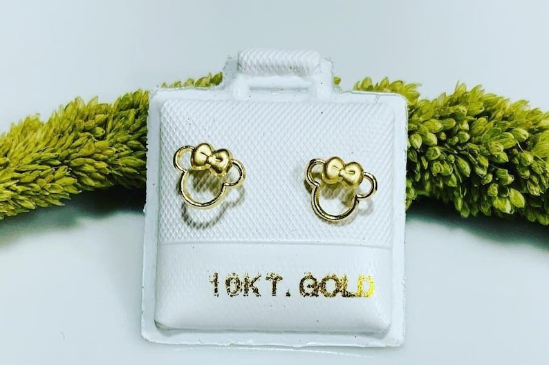 May include: A pair of gold-colored bow-shaped earrings on a white background. The earrings are in a small, white box with the text "10KT GOLD" printed on the bottom.