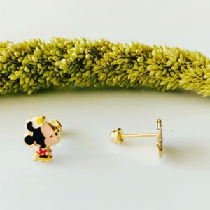 Puede incluir: Un par de pendientes de tono dorado con un personaje de dibujos animados con cabello negro, un lazo rojo y un sombrero amarillo. Los pendientes est&aacute;n sobre un fondo blanco.