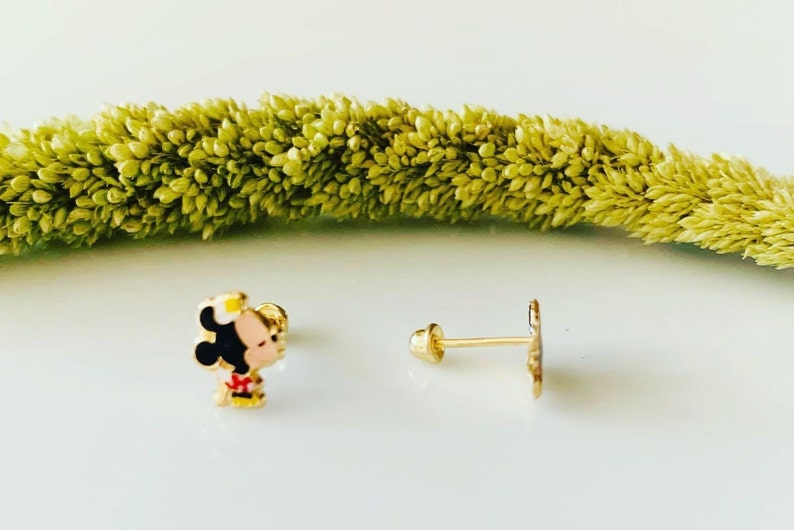 Puede incluir: Un par de pendientes de tono dorado con un personaje de dibujos animados con cabello negro, un lazo rojo y un sombrero amarillo. Los pendientes est&aacute;n sobre un fondo blanco.