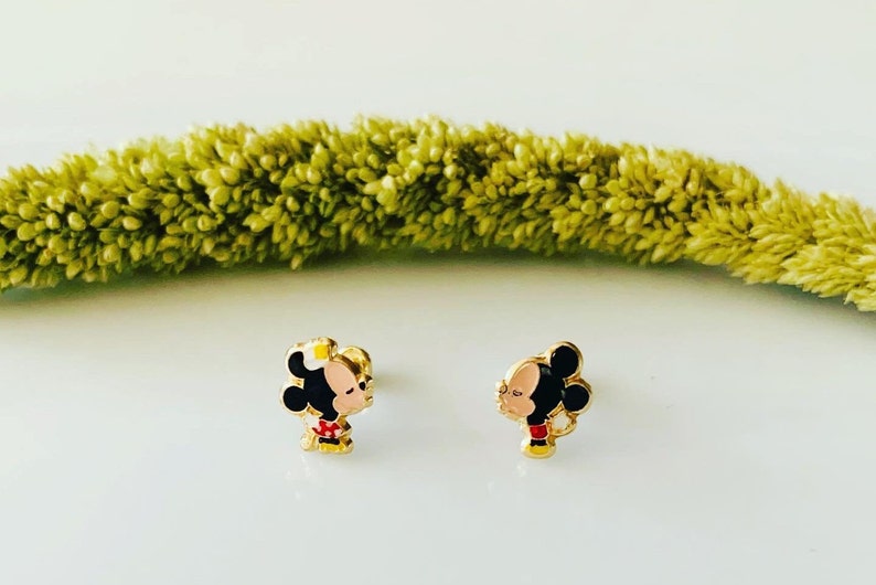 Puede incluir: Un par de pendientes de tono dorado con una imagen de dibujos animados de Mickey Mouse y Minnie Mouse bes&aacute;ndose. Los pendientes son peque&ntilde;os y delicados, perfectos para el uso diario.