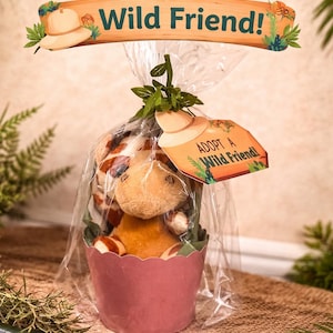 May include: A plush giraffe toy in a pink paper cup, wrapped in clear cellophane. The tag reads "Adopt a Wild Friend!" with a safari hat illustration. The top banner also says "Adopt a Wild Friend!"