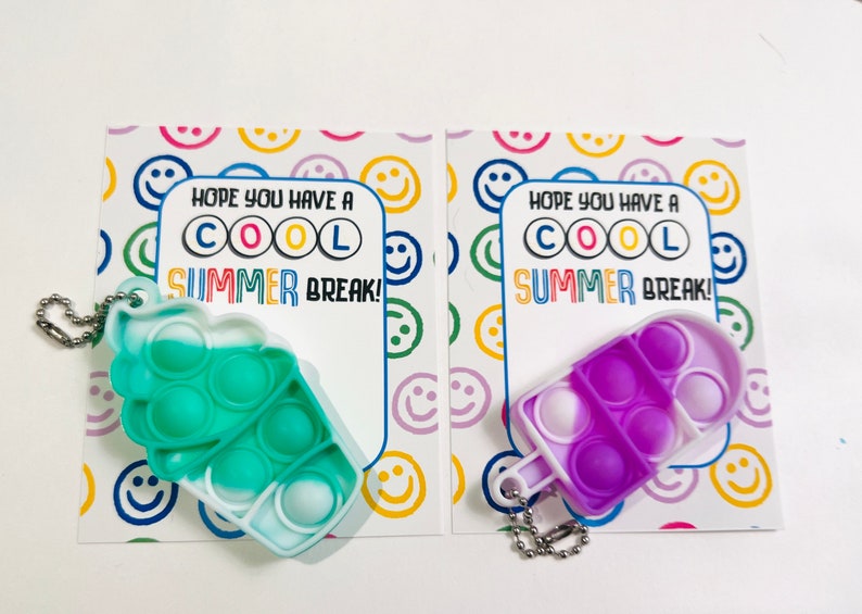 K&ouml;nnte beinhalten: Zwei bunte Pop-It-Fidget-Spielzeuge in Form von Eiswaffeln mit Schl&uuml;sselanh&auml;ngern. Die Spielzeuge sind gr&uuml;n und lila mit wei&szlig;en Akzenten. Jedes Spielzeug befindet sich neben einer Karte mit der Aufschrift "Ich hoffe, du hast coole Sommerferien!" mit einem Smiley-Muster im Hintergrund.